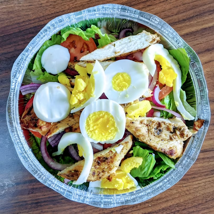 A BBQ chicken salad with hard-boiled eggs, tomatoes, and red onions in a foil container.