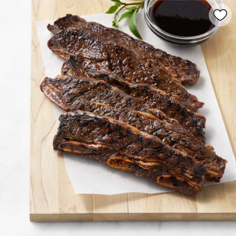 Grilled Riva's BBQ short ribs with a side of sauce and herbs on a wooden board.
