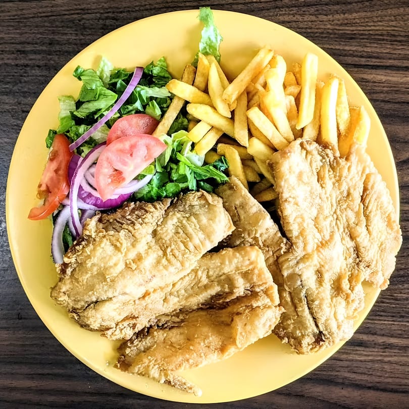 Fried fish fillets, french fries, and a fresh salad with tomatoes and red onion on a yellow plate.
